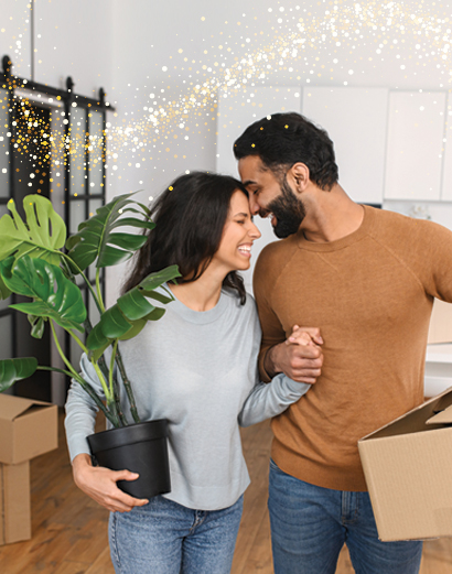 Couple moving into their new home. Young woman holding plant while guy is holding a box embracing each other with smiles.
