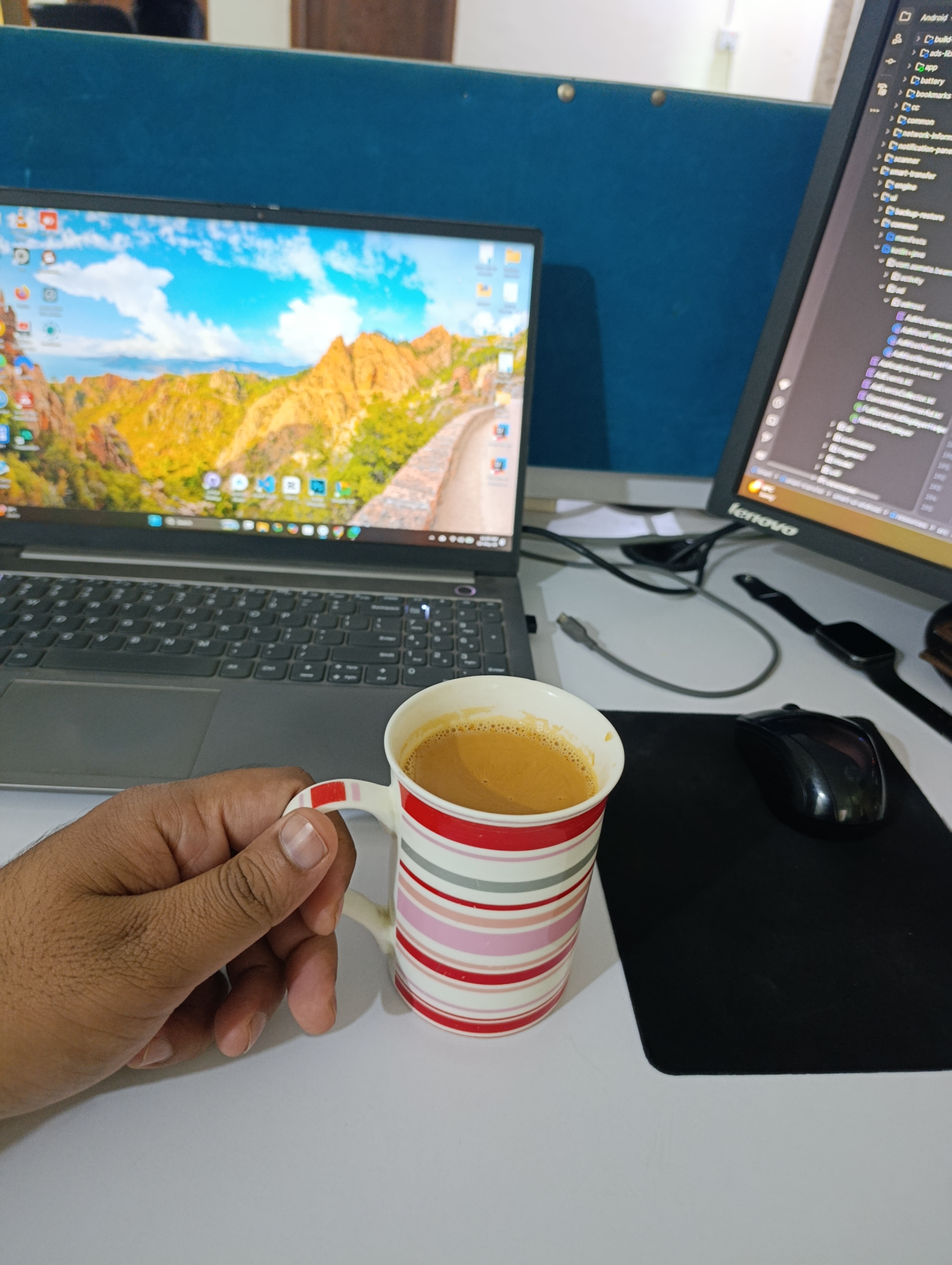 A hand holds a striped mug of tea at a white desk with a laptop and monitor, creating a focused workspace.